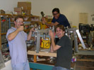 Mark hacksawing at a leg of the robot, Eiki threatening to cut wires, and Sheraz standing above, wielding a large wrench as though to smash the robot